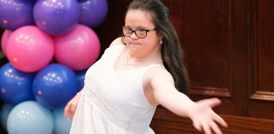 A young girl with Down Syndrome dances, with a display of balloons in the background