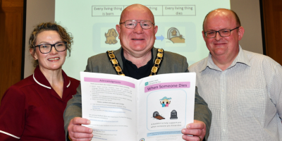 A woman and two men standing beside each other. One is wearing a maroon tunic, and the man in the middle is holding a booklet and wearing a mayor's chain.