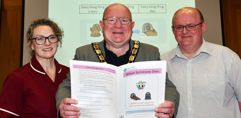 A woman and two men standing beside each other. One is wearing a maroon tunic, and the man in the middle is holding a booklet and wearing a mayor's chain.
