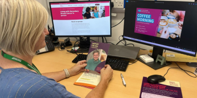 A woman sits at a desk with two computer screens in front of her. She is looking at information about Breast Cancer Now.
