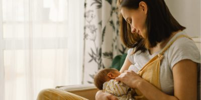 A breastfeeding mother with her baby, sitting by a window with a white curtain.