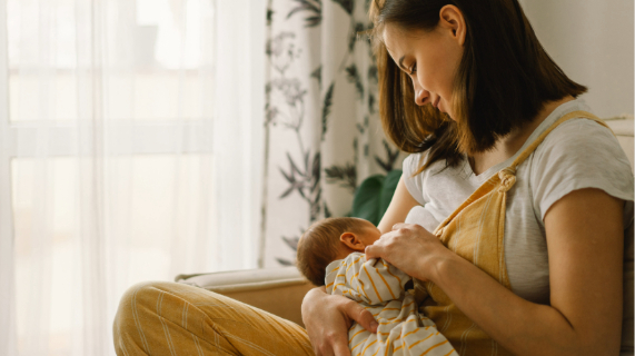A breastfeeding mother with her baby, sitting by a window with a white curtain.