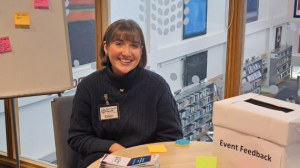 A woman sits at a desk. There are posy0it notes on the desk and a box for leaving feedback inside.