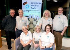 "A group of people standing and sitting beside a banner about Causeway Hospital Radio.