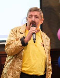 A man sings into a microphone, while wearing a gold sparkly jacket.