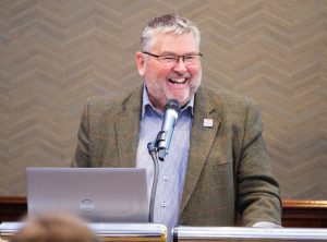 A man laughs while standing at a podium with a microphone and laptop in front of him.