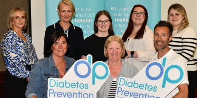 Eight people, some sitting and some standing. Those sitting at the front are holding signs for the Diabetes Prevention Programme.