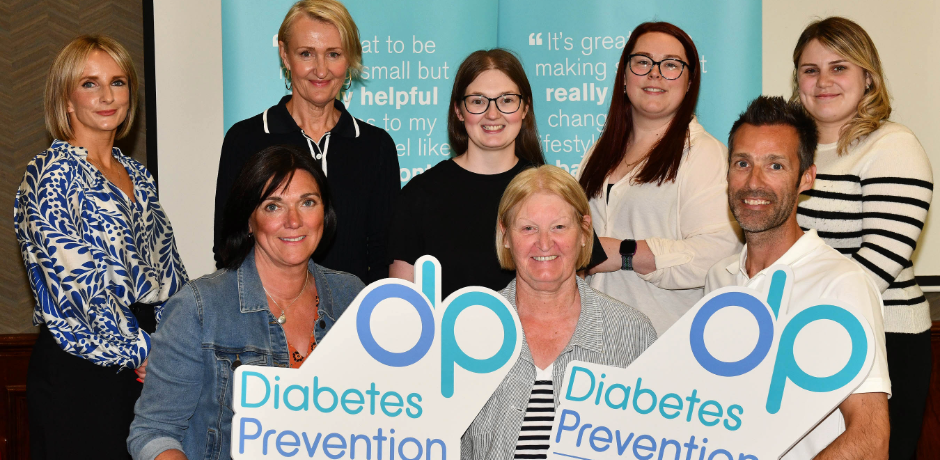 Eight people, some sitting and some standing. Those sitting at the front are holding signs for the Diabetes Prevention Programme.