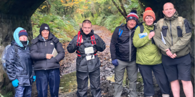 A group of people wearing outdoor clothes standing under a bridge in a forest.