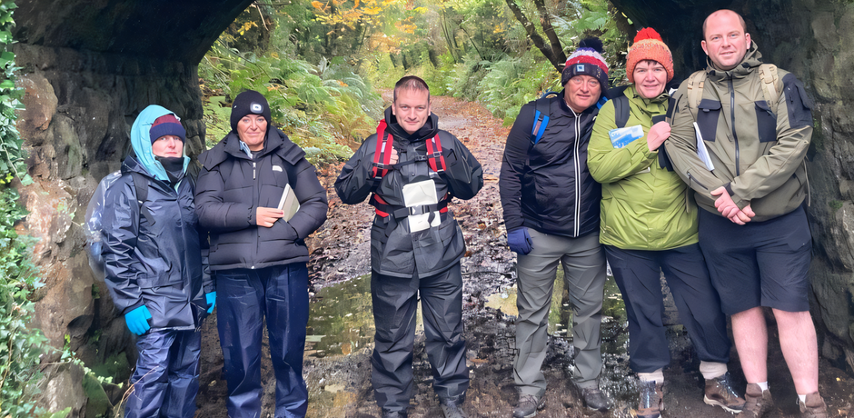 A group of people wearing outdoor clothes standing under a bridge in a forest.