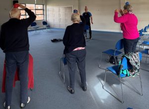 Participants in a strength and balance class, standing beside chairs while balancing a small ball on their head.