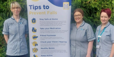 Three healthcare workers wearing grey uniform tunics, standing beside a pop-up featuring information about falls prevention.