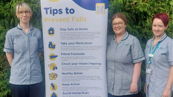 Three healthcare workers wearing grey uniform tunics, standing beside a pop-up featuring information about falls prevention.
