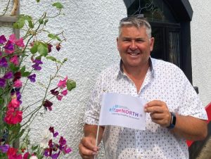 Garth Anderson standing outside holding a small white flag with Team North branding on it. A white building and purple and pink flower are in the background.