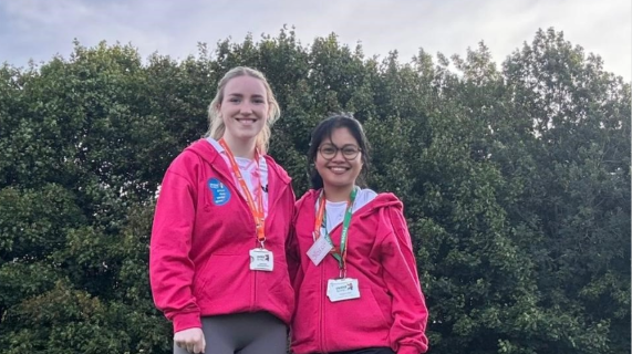 Hazelle Ongcoy and Caillee McHugh standing together outside. They are wearing matching pink jumpers and there are trees behind them.