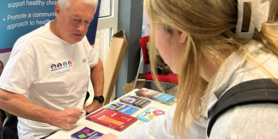 A volunteer wearing a Northern Trust branded t-short sits behind an information table filled with literature. A young woman is leaning over to pick something up off the table.
