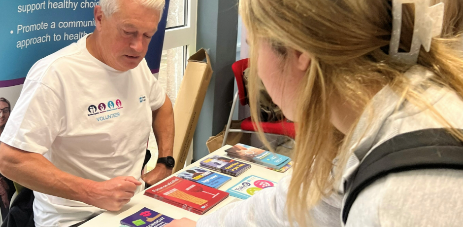 A volunteer wearing a Northern Trust branded t-short sits behind an information table filled with literature. A young woman is leaning over to pick something up off the table.