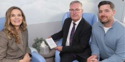 The Health Minister holds a framed poem while sitting with Corrina and Johnny McAleese in the Infinity Room.