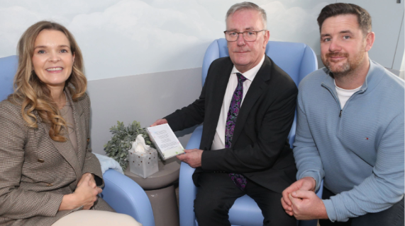 The Health Minister holds a framed poem while sitting with Corrina and Johnny McAleese in the Infinity Room.