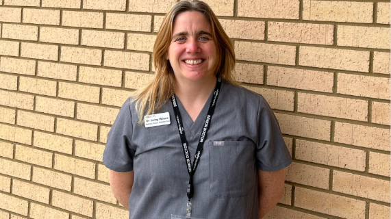 Dr Jennifer Wilson wearing a grey uniform standing against a brick wall.