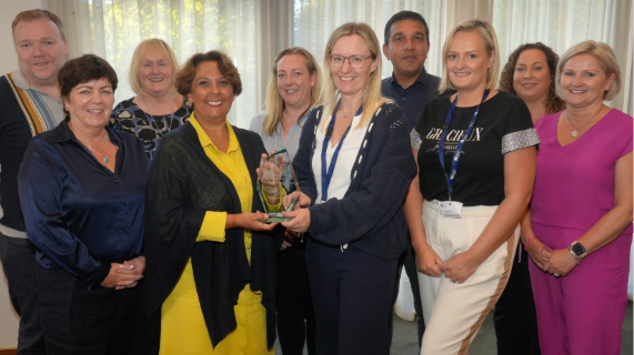 Ten people standing together. One lady in the group is receiving an award from another lady.