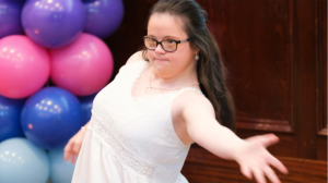 A young girl with Down Syndrome dances, with a display of balloons in the background