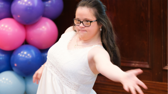 A young girl with Down Syndrome dances, with a display of balloons in the background