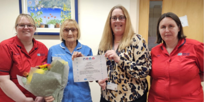 Four women stand together. Two are wearing red tunics. One is holding a bouquet of flowers and a certificate.