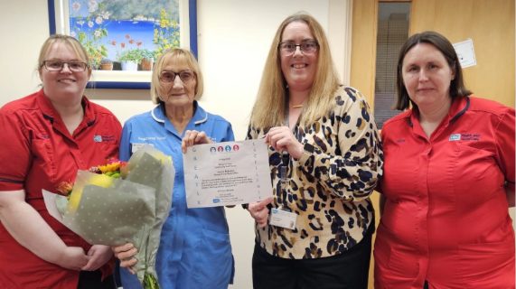Four women stand together. Two are wearing red tunics. One is holding a bouquet of flowers and a certificate.