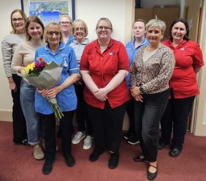 Nine women standing together. Some are wearing healthcare tunics. One is holding a bouquet of flowers.