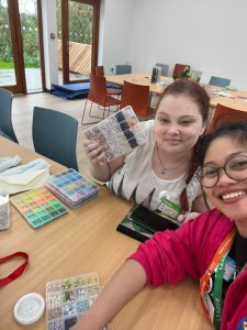 Hazelle sits beside a young person at a table. The young person is holding a box of beads, while there are more boxes on the table.