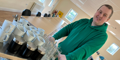 A man stands beside as trolley filled with cups, with tables behind him.