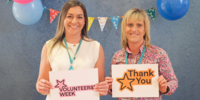 Two women standing beside each other with bunting and balloons in the background, One is holding a sign which says 'Volunteers Week' and the other is holding a sign which says 'thank you'