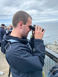 A service user stands at a railing looking out to sea through binoculars.