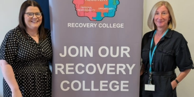 Two women stand beside a pop-up about banner with text on it which reads 'join our recovery college'.