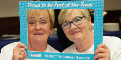 Two women looking through a selfie frame with text on it which reads 'proud to be part of the team', NHSCT volunteer service.