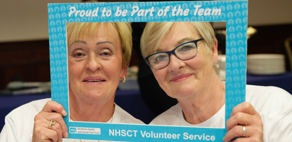 Two women looking through a selfie frame with text on it which reads 'proud to be part of the team', NHSCT volunteer service.