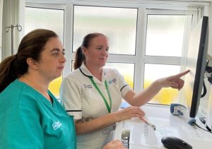Two people look at a screen together. One is wearing a green tunic and the other one is wearing a white tunic.
