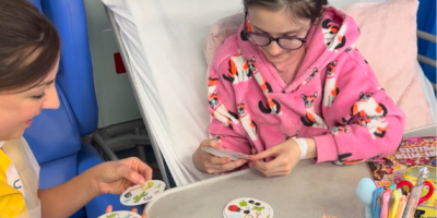 A Play Specialist wearing a yellow tunic sits beside a young patient's bed. The patient is sitting up in bed and the two are playing a card game together.