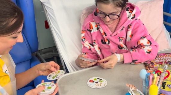 A Play Specialist wearing a yellow tunic sits beside a young patient's bed. The patient is sitting up in bed and the two are playing a card game together.