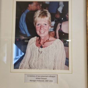 A framed photograph of a woman. Text reads 'in memory of our esteemed colleague Arlene Stewart, Manager Hollybank 1987-2016.