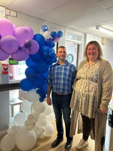 A man and woman stand beside a balloon arch.