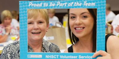 Two women smiling through a selfie frame with text on it which reads 'proud to be part of the tean, NHSCT volunteer service'.