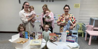 Three mums holding their babies with two toddlers standing beside them. Two tables are in front, with buns, cakes and information leaflets.