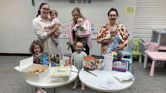 Three mums holding their babies with two toddlers standing beside them. Two tables are in front, with buns, cakes and information leaflets.