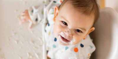 A young baby looks up at the camera while sitting in a high chair with food on his tray and face.