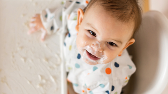 A young baby looks up at the camera while sitting in a high chair with food on his tray and face.