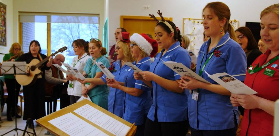 Hospital staff at a carol service. Some are wearing festive hair band and one is playing a guitar.