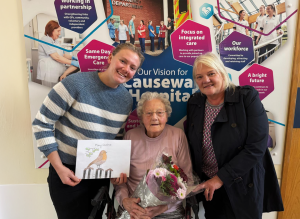 Ethel, winner of the Christmas card competition, sits in a chair holding a bunch of flowers, along with her daughter and granddaughter. Her granddaughter is holding a copy of her winning card design which shows a robin on a branch.