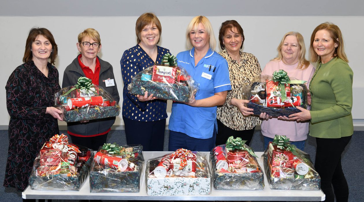 Seven women standing behind a table. They are holding hampers wrapped in festive wrapping. There are more hampers sitting on the table.